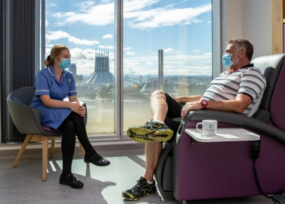 Picture showing a nurse sitting with a patient who is receiving chemotherapy. The Liverpool skyline is in the distance behind them